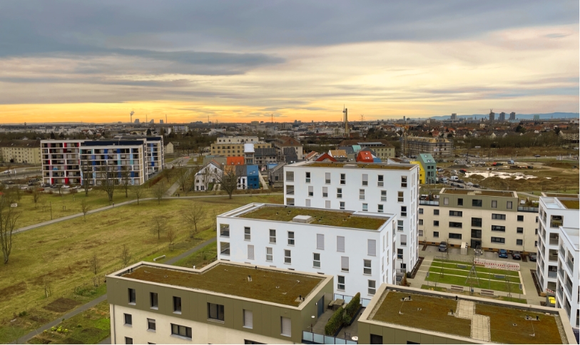 KI-basierte Bildbeschreibung: Eine Stadtlandschaft bei Sonnenuntergang mit modernen weißen Wohnhäusern mit grünen Dächern im Vordergrund, grasbewachsenen Freiflächen und bunten älteren Gebäuden im Hintergrund unter einem bewölkten Himmel.