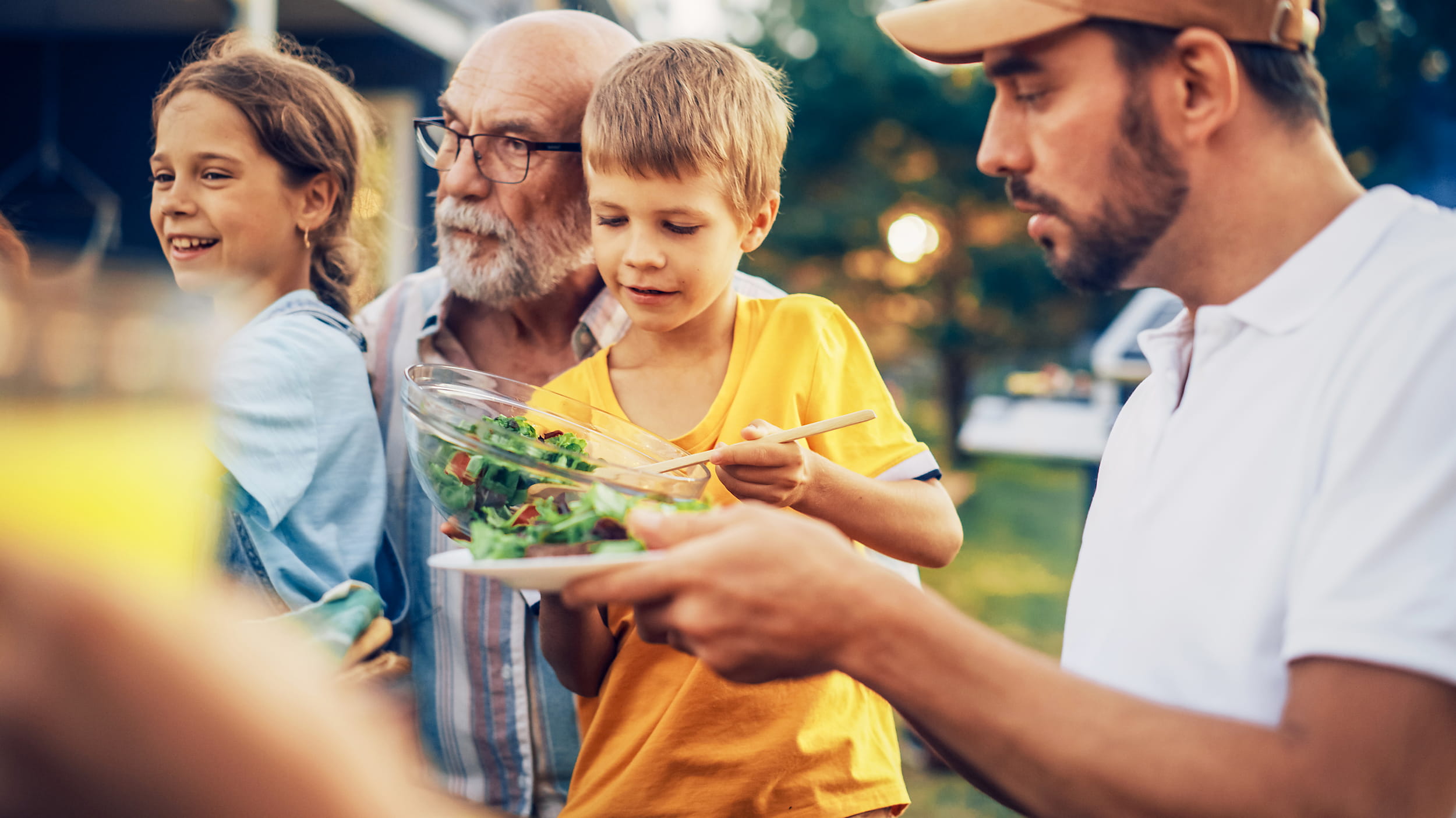 KI-basierte Bildbeschreibung: Ein kleiner Junge serviert Salat auf seinem Teller, umgeben von Familienmitgliedern im Freien. Ein älterer Mann und ein Mädchen schauen zu, während ein Mann mit einer Mütze in der Nähe steht. Sie alle scheinen eine gemeinsame Mahlzeit in einem Hinterhof zu genießen.