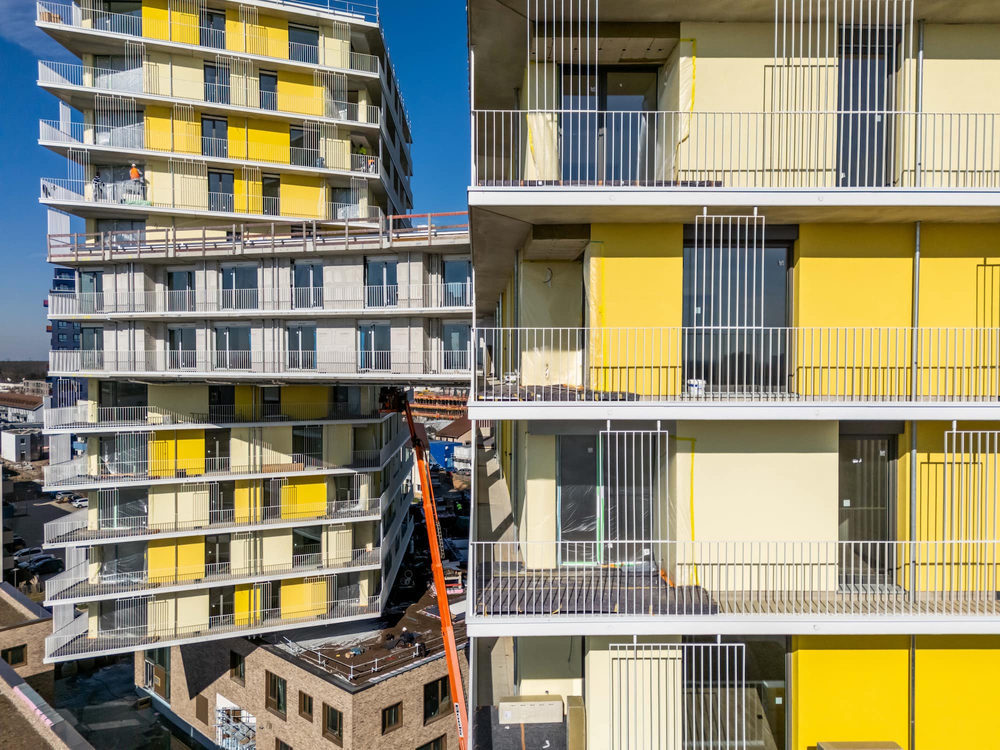 KI-basierte Bildbeschreibung: Moderne Wohngebäude mit gelben Balkonen und weißen Geländern; ein Gebäude ist gekippt und mit einem anderen durch eine Hochbrücke verbunden. Bauarbeiter und Baumaschinen sind zu sehen. Heller, sonniger Tag mit einem klaren blauen Himmel.