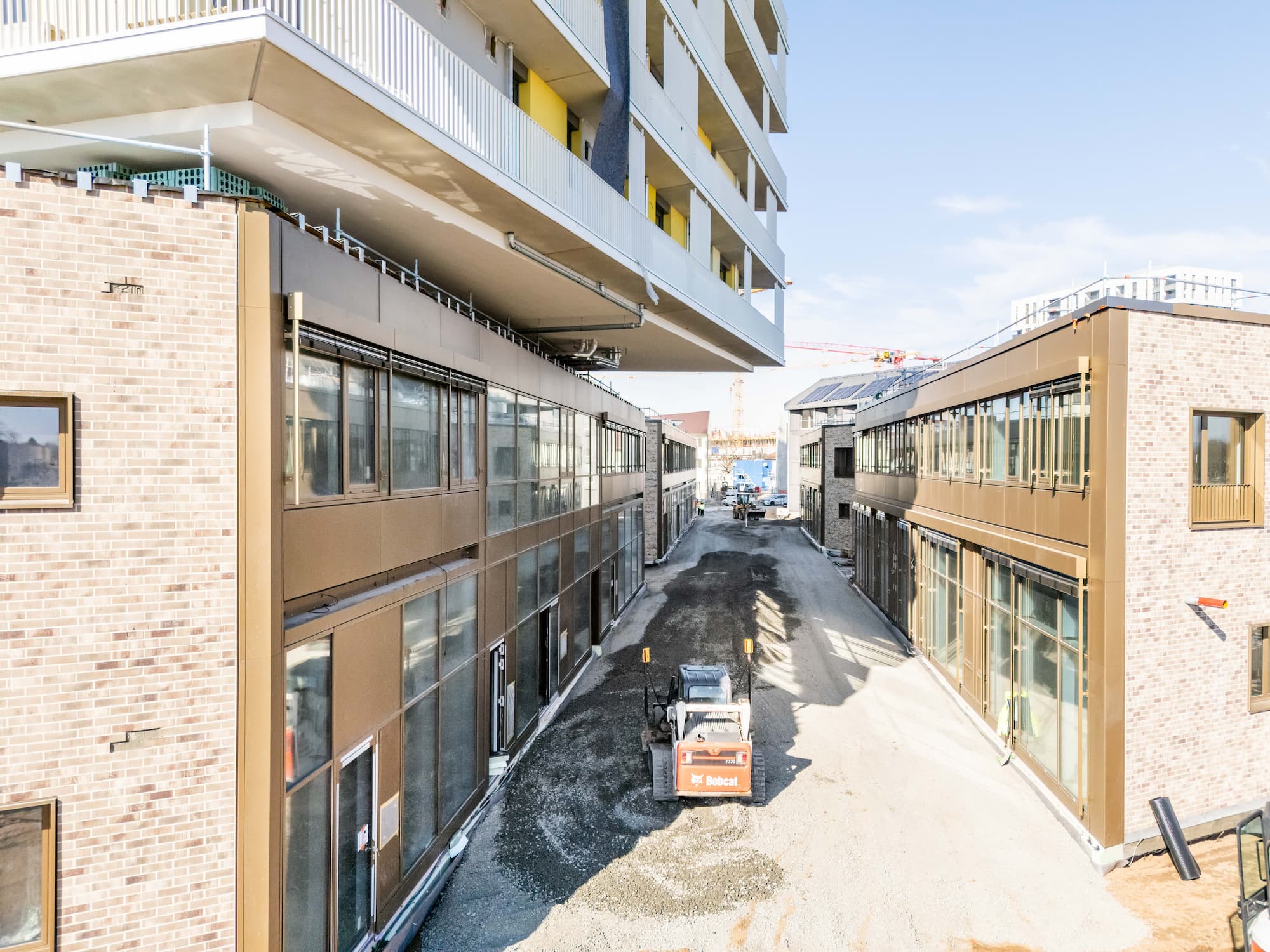 KI-basierte Bildbeschreibung: Eine Baustelle zwischen zwei modernen, mehrstöckigen Gebäuden mit Backsteinfassade. Ein kleines Baufahrzeug streut Schotter auf die Straße, und Baumaterialien liegen verstreut herum. Der Himmel ist klar.