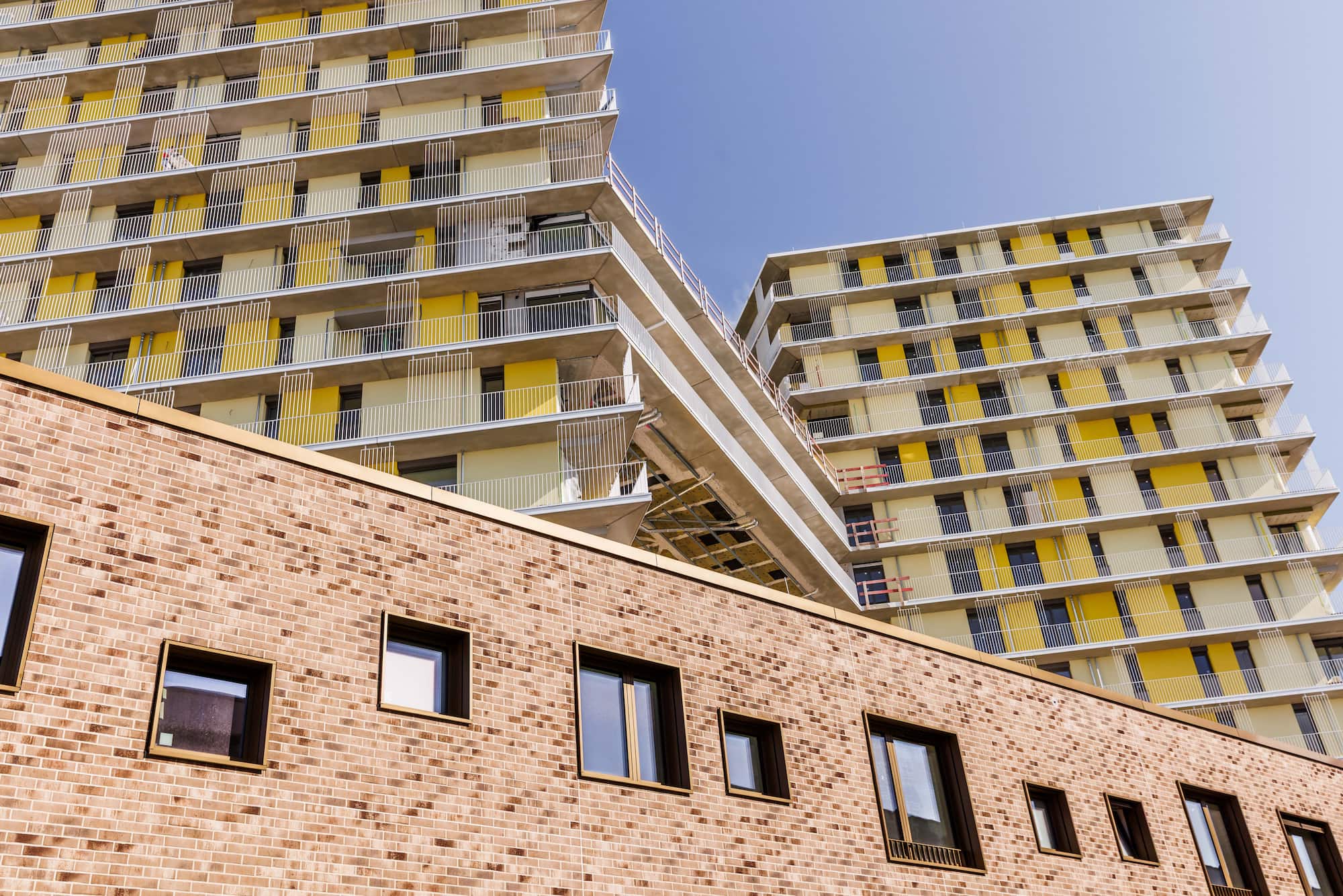 KI-basierte Bildbeschreibung: Hinter einer Backsteinfassade mit rechteckigen Fenstern erheben sich moderne Wohnhäuser mit gelben und weißen Balkonen vor einem strahlend blauen Himmel.