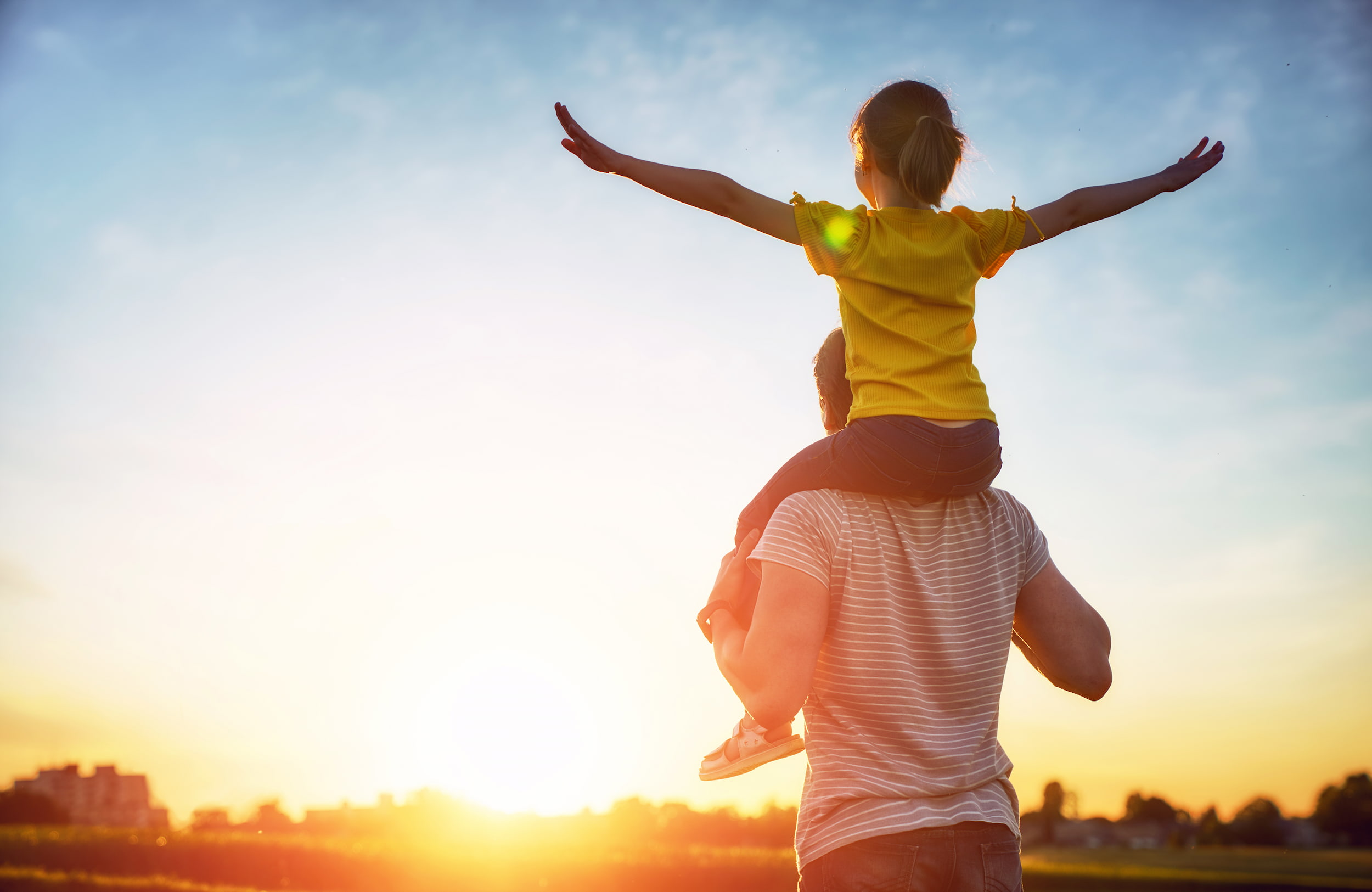 KI-basierte Bildbeschreibung: Ein Mann trägt bei Sonnenuntergang ein Kind auf seinen Schultern. Das Kind, das ein gelbes Hemd trägt, breitet die Arme aus, während beide der Sonne zugewandt sind, was eine warme, fröhliche Szene vor einem klaren Himmel ergibt.