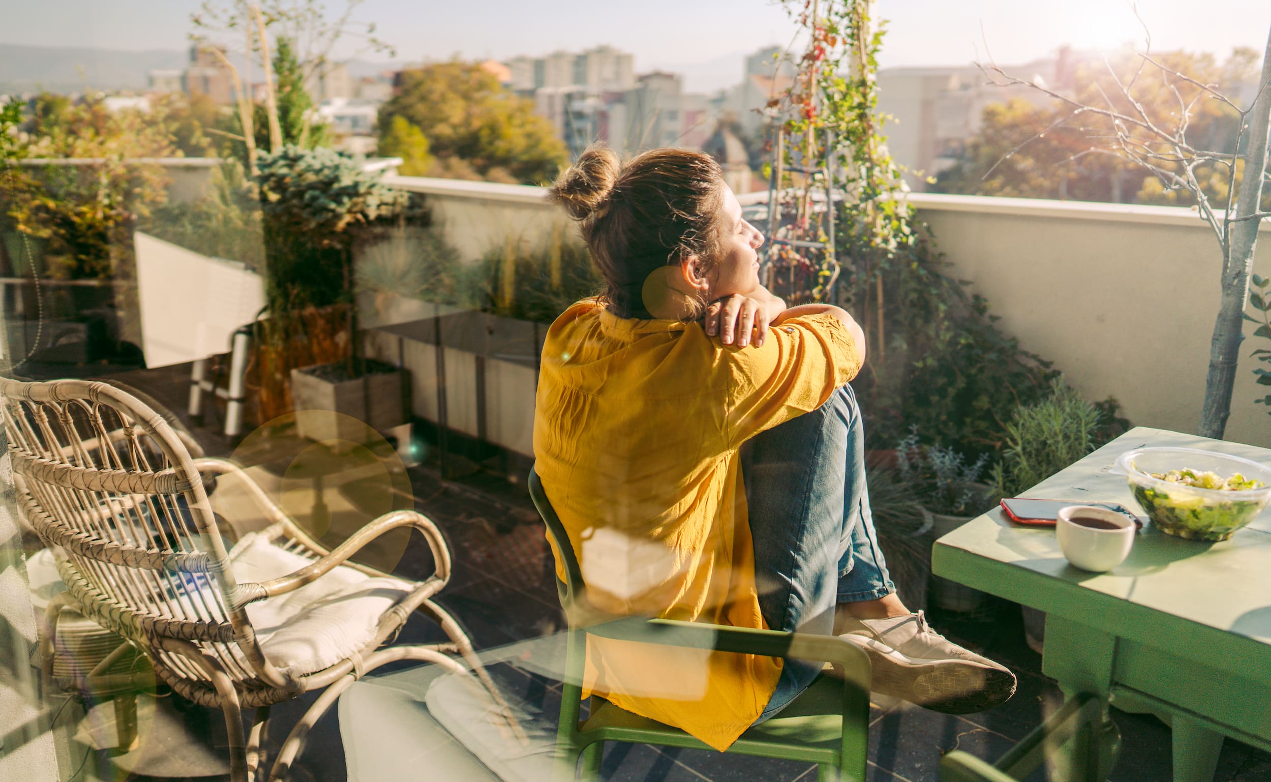 KI-basierte Bildbeschreibung: Eine Frau in einem gelben Hemd sitzt auf einem Balkon, stützt sich auf die Knie und blickt nachdenklich in die Ferne. Neben ihr stehen ein Tisch mit einer Salatschüssel und einem Kaffeebecher, ein Korbstuhl und die Stadtansicht im Hintergrund.