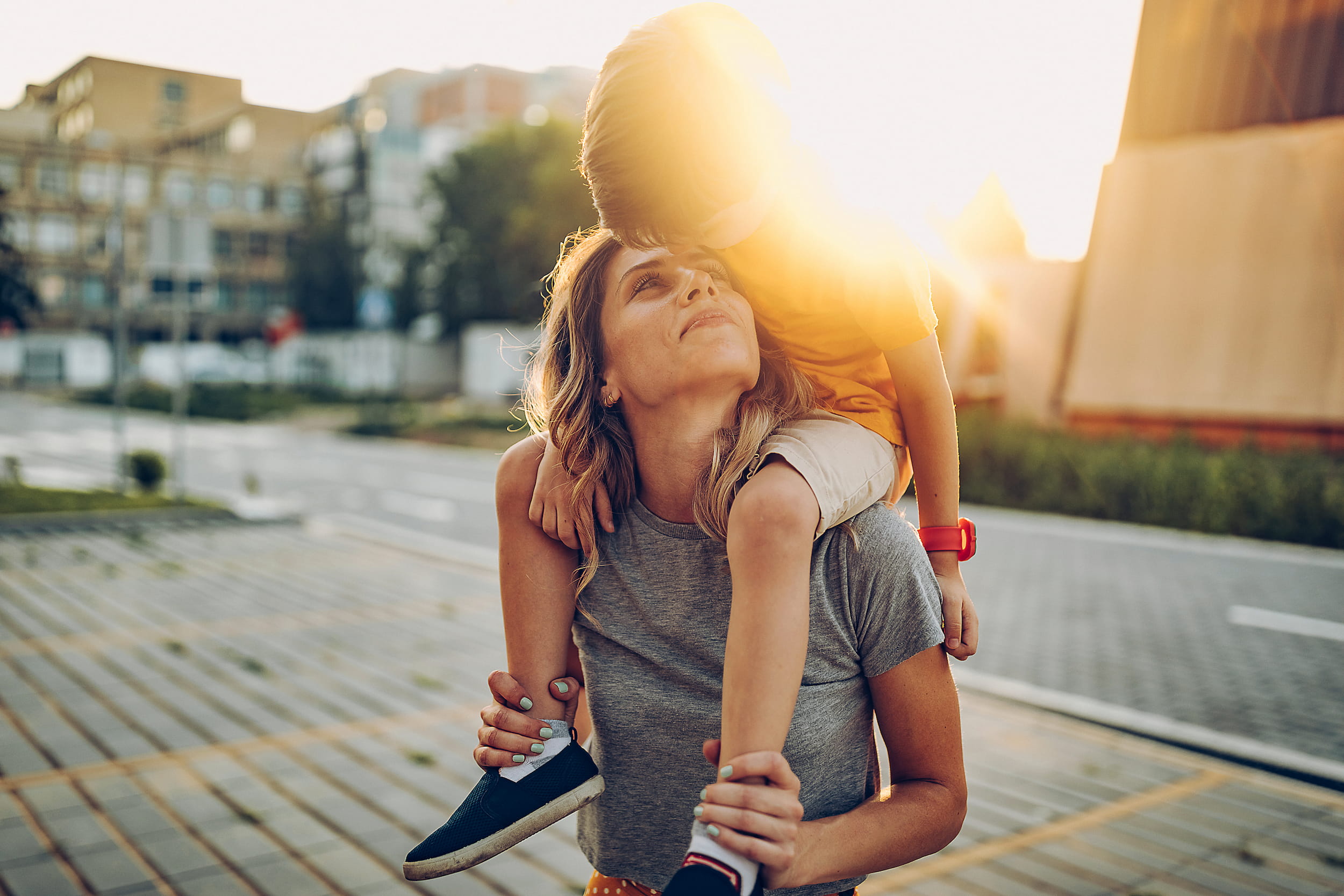 KI-basierte Bildbeschreibung: Eine Frau trägt ein Kind auf ihren Schultern im Freien bei Sonnenuntergang. Das Kind beugt sich zu ihr herunter, um sie auf die Stirn zu küssen, und das Sonnenlicht scheint hell hinter ihnen. Sie wirken glücklich und entspannt in einer städtischen Umgebung.