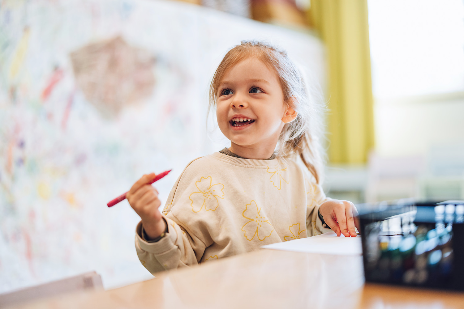 KI-basierte Bildbeschreibung: Ein junges Mädchen lächelt, während es einen roten Buntstift hält und an einem Tisch mit Papier und einem schwarzen Behälter sitzt. Der Hintergrund zeigt eine bunte, bekritzelte Wand, die eine spielerische, kreative Umgebung suggeriert.