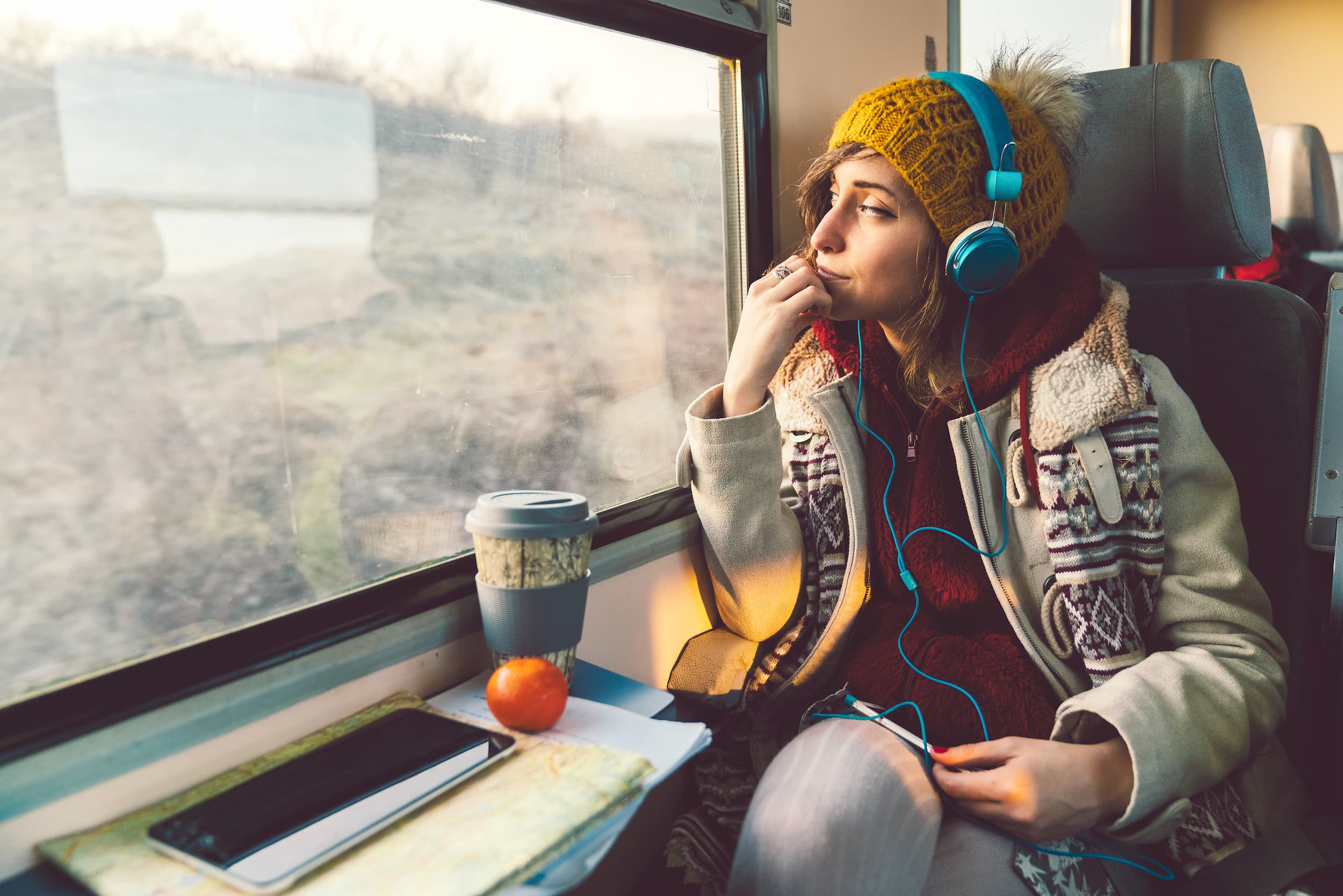 KI-basierte Bildbeschreibung: Eine Frau mit gelber Strickmütze und blauen Kopfhörern schaut nachdenklich aus dem Fenster eines Zuges. Sie sitzt mit einer Kaffeetasse, einer Karte, einem Apfel und ihrem Telefon auf dem Tisch vor ihr.