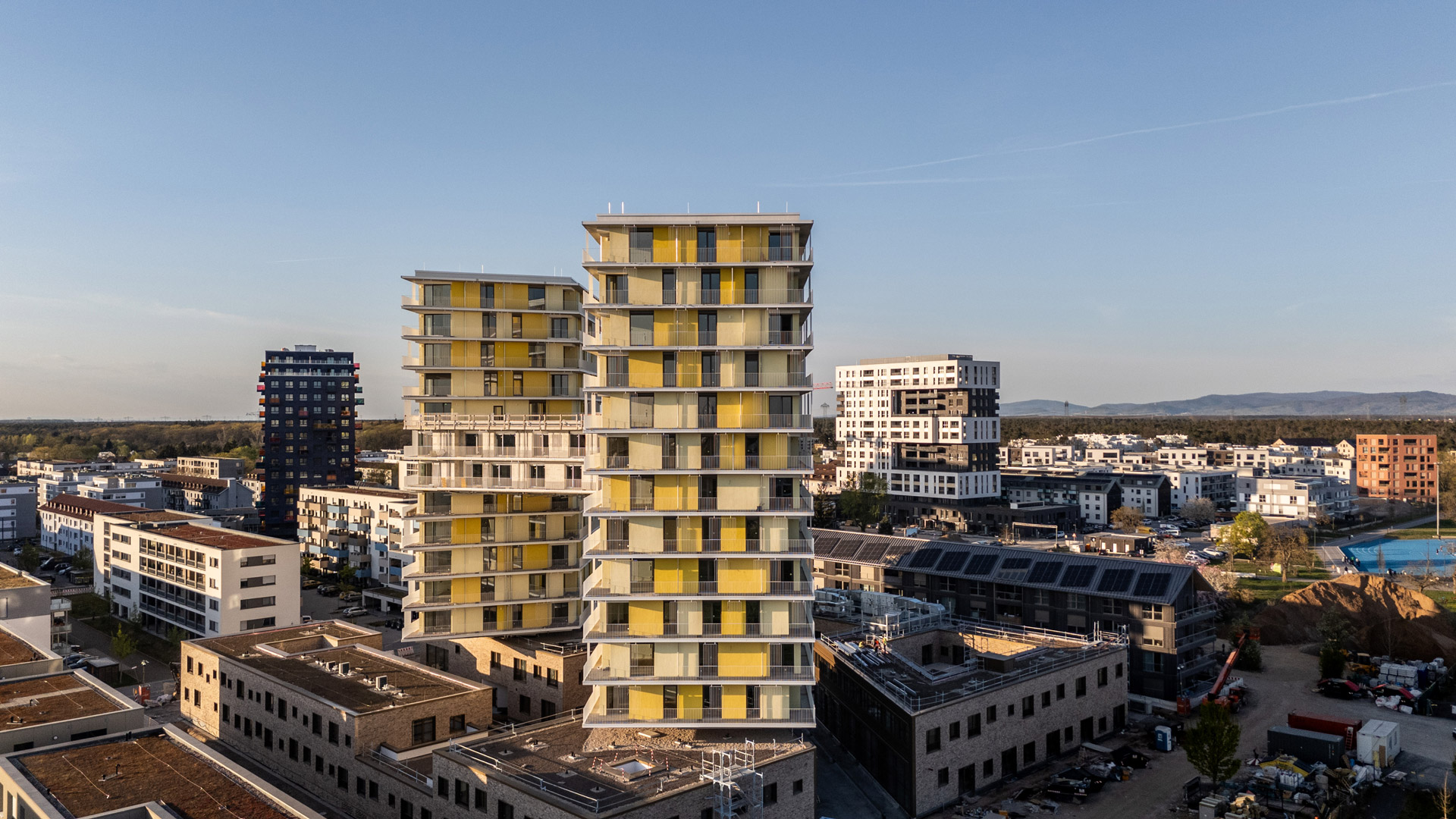 KI-basierte Bildbeschreibung: Luftaufnahme von modernen städtischen Gebäuden mit zwei hohen Türmen mit gelben und weißen Fassadenplatten, umgeben von anderen Wohn- und Geschäftsgebäuden unter klarem Himmel.