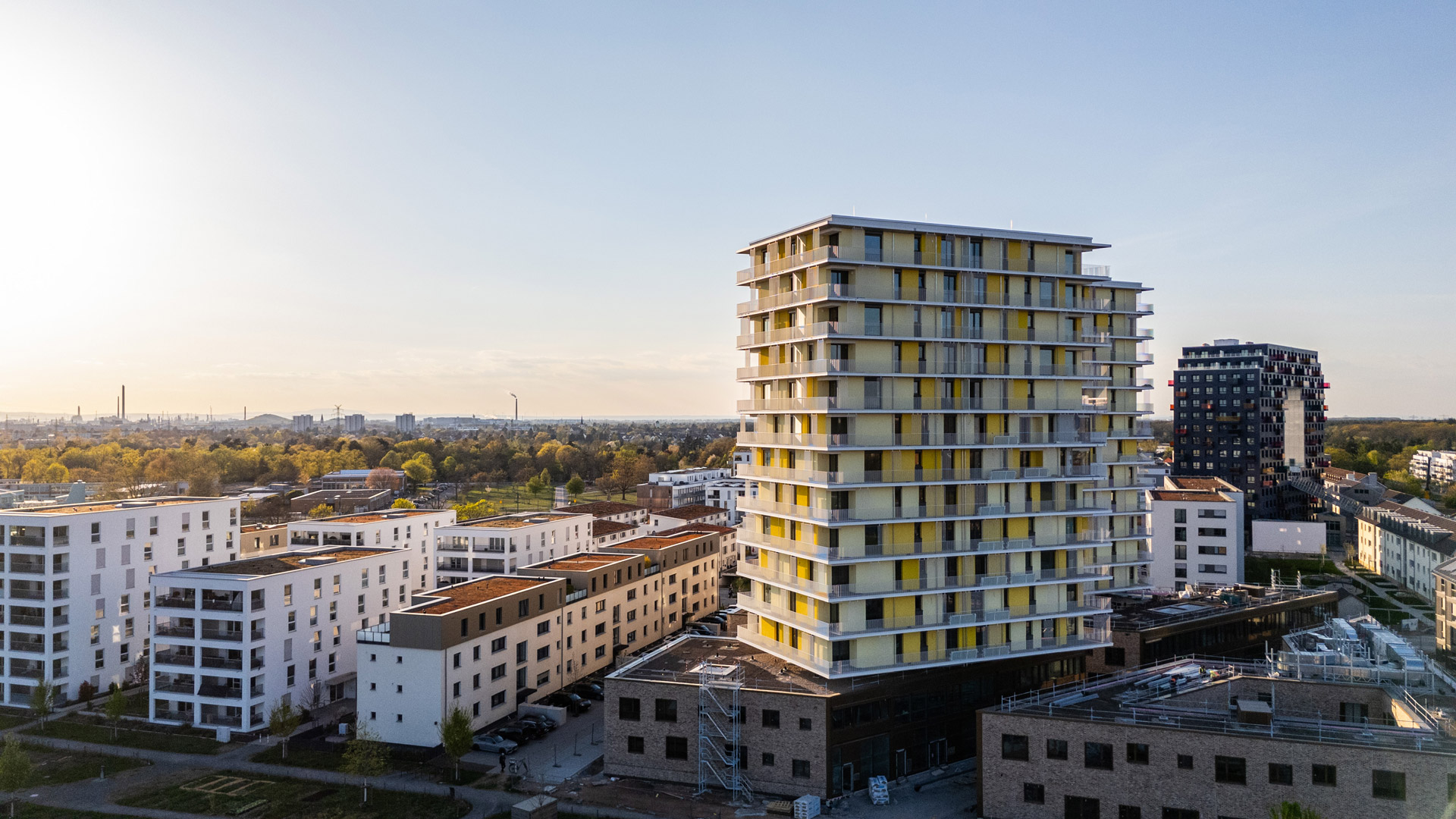 KI-basierte Bildbeschreibung: Ein modernes Stadtviertel mit mittelhohen Wohnhäusern, darunter ein zentrales Hochhaus mit gelben Akzenten, fotografiert bei Sonnenuntergang unter klarem Himmel.