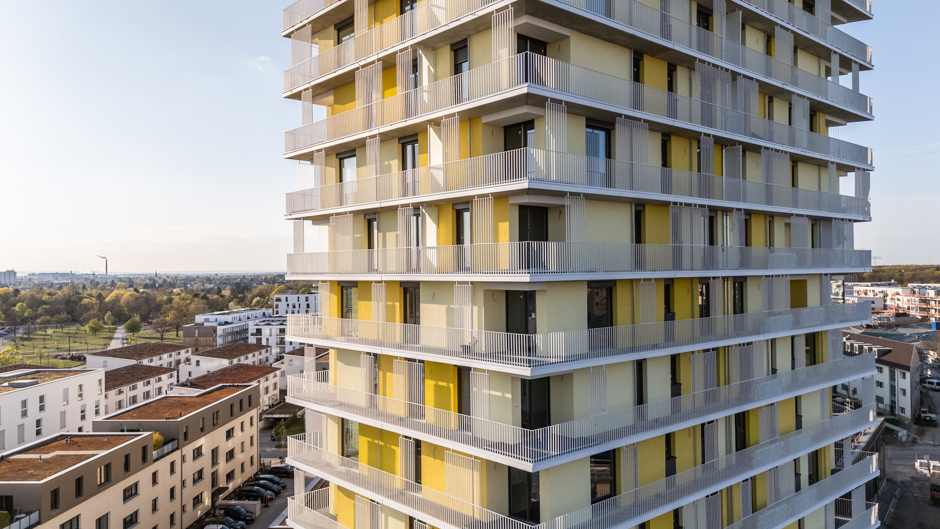 KI-basierte Bildbeschreibung: Modernes Hochhaus mit gelben und beigen Paneelen und umlaufenden Balkonen, umgeben von niedrigeren Wohngebäuden und Grünanlagen unter einem klaren Himmel.