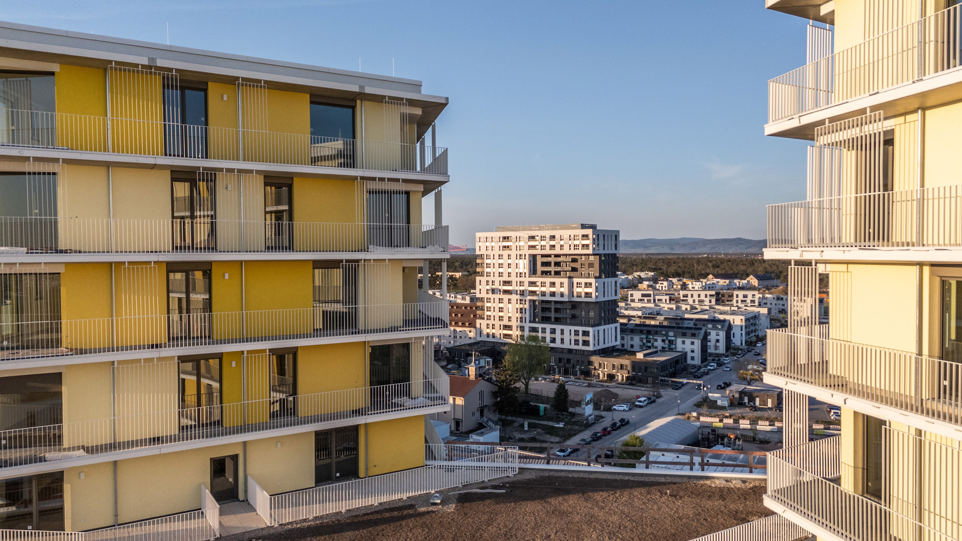 KI-basierte Bildbeschreibung: Zwei moderne Mehrfamilienhäuser mit gelben Akzenten umrahmen die Ansicht eines Stadtgebiets mit weiteren Wohngebäuden, Straßen und Bäumen unter einem klaren blauen Himmel.