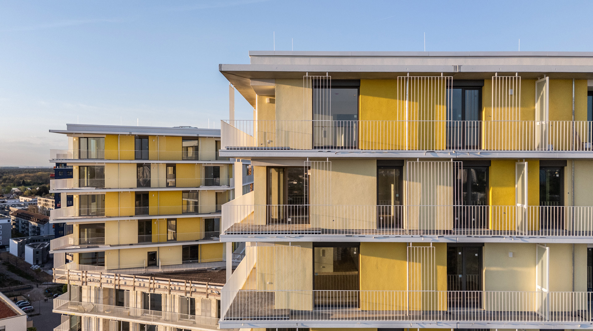 KI-basierte Bildbeschreibung: Moderne Mehrfamilienhäuser mit gelber und weißer Außenverkleidung, großen Fenstern und Balkonen mit Metallgeländern vor einem klaren blauen Himmel. Das Sonnenlicht wirft Schatten auf die Fassaden.