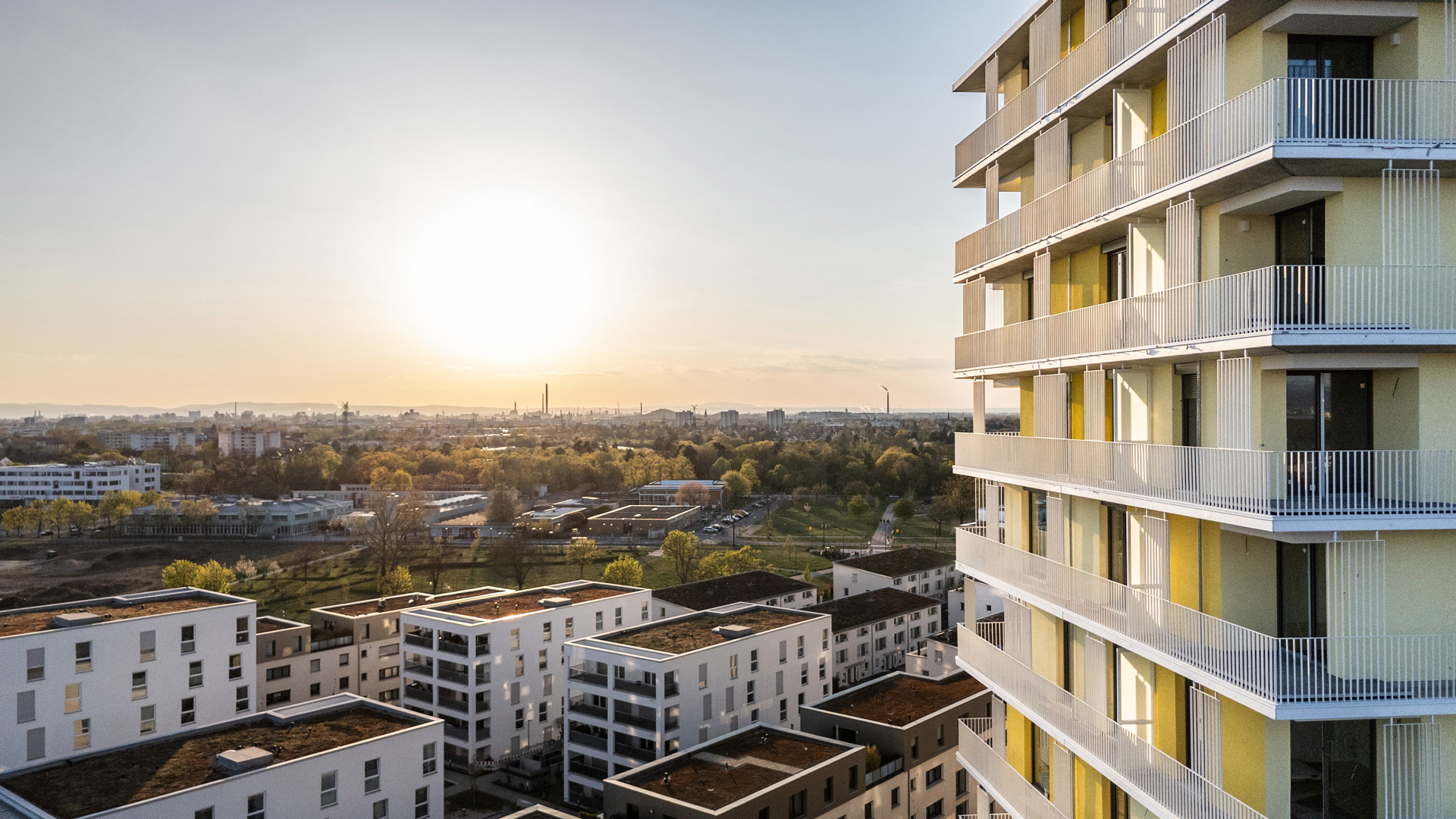 KI-basierte Bildbeschreibung: Im Vordergrund ist ein modernes Mehrfamilienhaus mit Balkonen bei Sonnenuntergang zu sehen, mit Blick auf mehrere andere Wohngebäude und eine Stadtlandschaft mit Bäumen und Freiflächen.