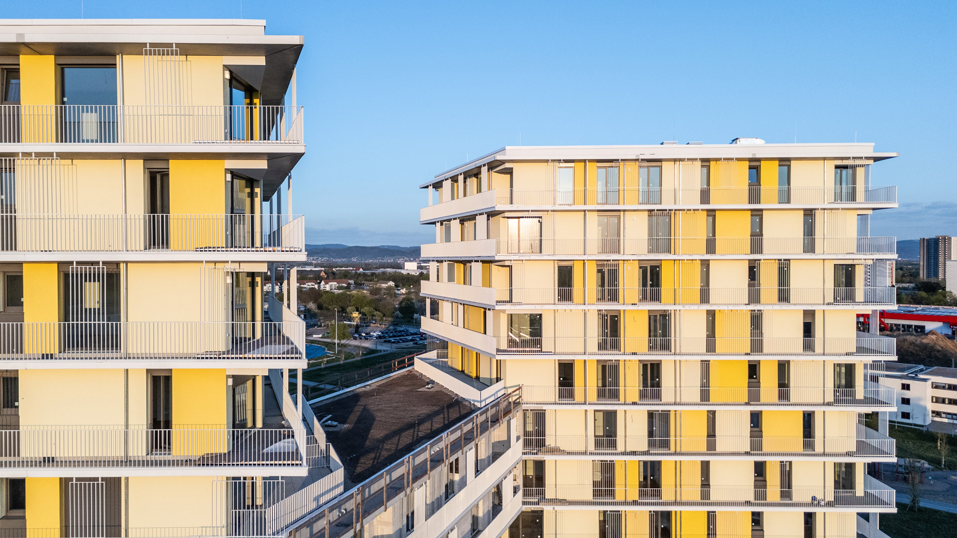 KI-basierte Bildbeschreibung: Zwei moderne Wohngebäude mit gelben und weißen Fassaden, großen Fenstern und Balkonen, die durch einen Gehweg miteinander verbunden sind, aufgenommen bei Tageslicht mit einem klaren Himmel und einer Stadtlandschaft im Hintergrund.