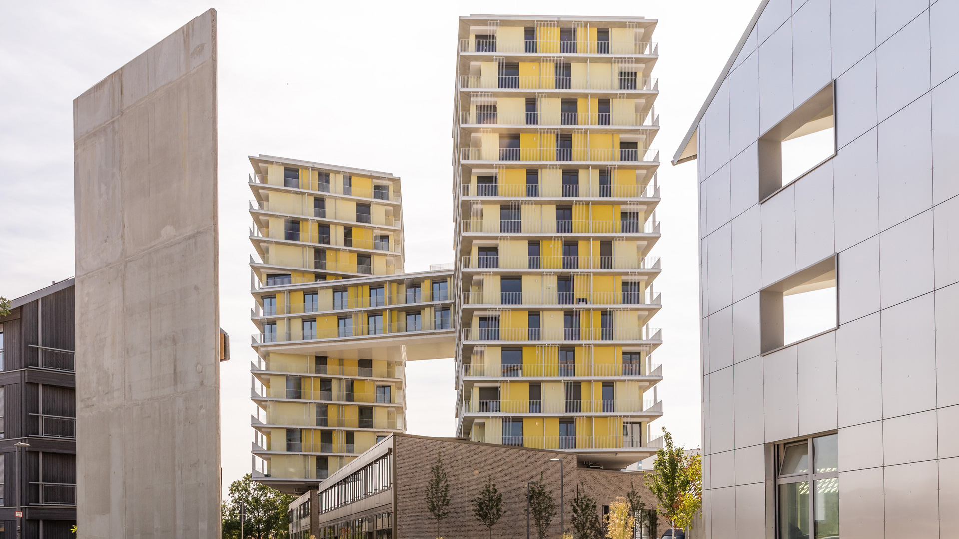 KI-basierte Bildbeschreibung: Zwei moderne Hochhäuser mit gelben und weißen Balkonen sind durch eine Skybridge verbunden. Im Vordergrund befindet sich links eine Betonmauer und rechts ein weißes Gebäude mit quadratischen Fenstern.