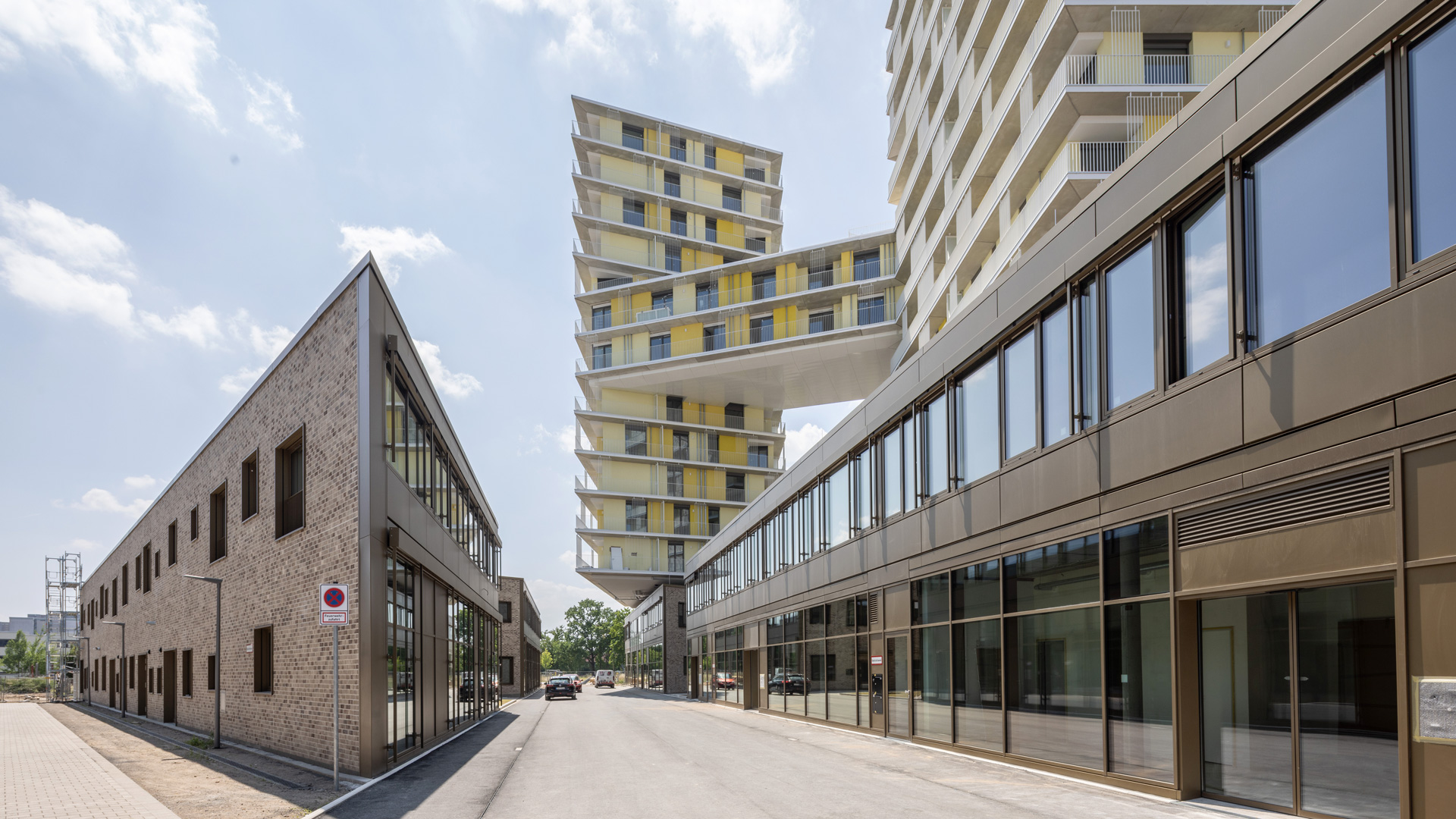 KI-basierte Bildbeschreibung: Eine moderne Straße mit kantigen Gebäuden; rechts ein Hochhaus mit gestapelten, unregelmäßig angeordneten Stockwerken und einer zentralen Brücke, links ein Backsteinbau mit großen Fenstern unter blauem Himmel.