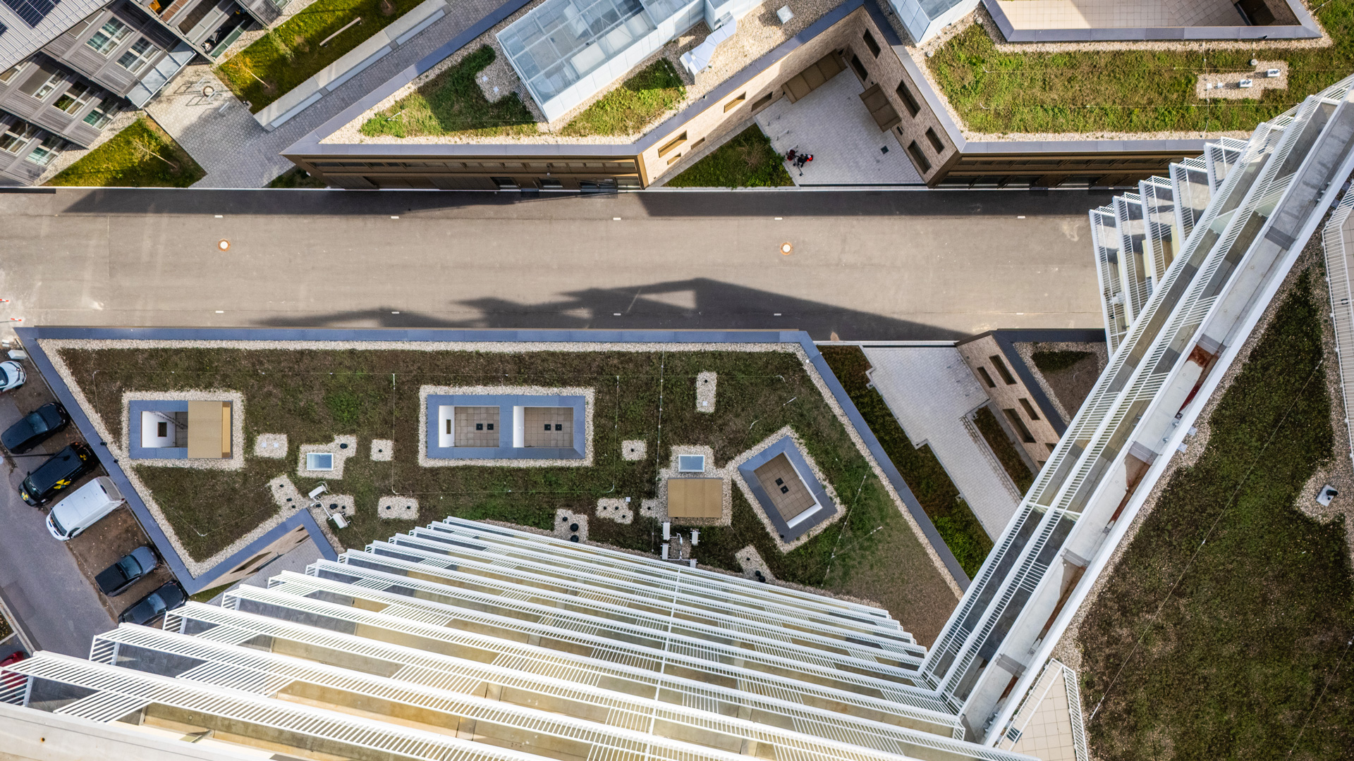 KI-basierte Bildbeschreibung: Luftaufnahme von modernen städtischen Gebäuden mit geometrischen Formen, grünen Dächern, einer Straße, die durch das Zentrum verläuft, und geparkten Autos an der Seite. Das Bild wurde von einem hohen Gebäude aus aufgenommen, das gerade nach unten schaut.