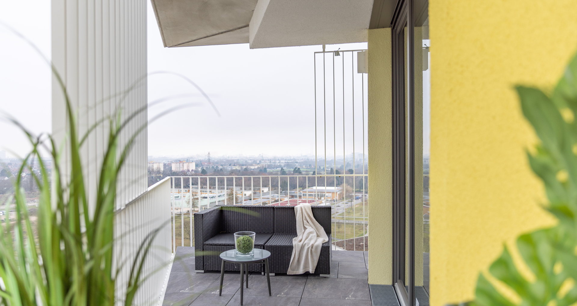 KI-basierte Bildbeschreibung: Ein moderner Balkon mit einem schwarzen Outdoor-Sofa, einem kleinen Glastisch mit einer Topfpflanze und einer beigen Decke, die über das Sofa drapiert ist. Der Balkon bietet einen Blick auf eine Stadtlandschaft unter einem bewölkten Himmel, mit Grün im Vordergrund.