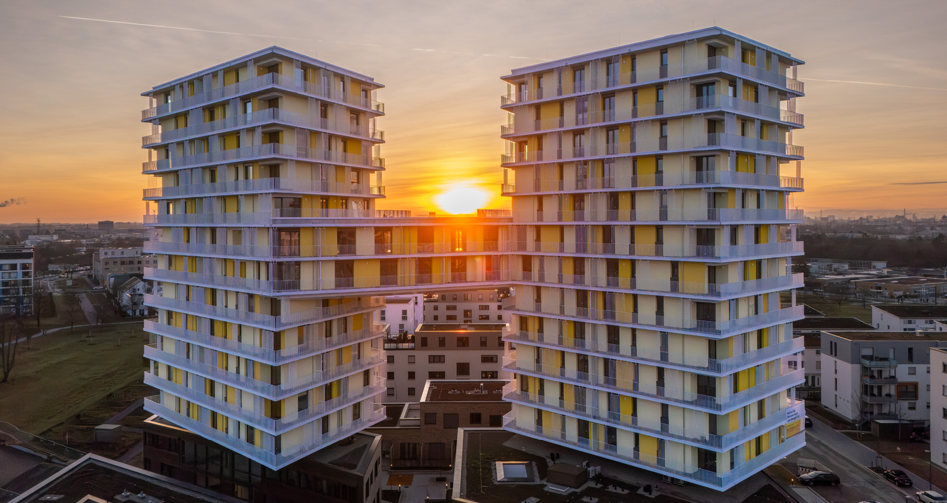 KI-basierte Bildbeschreibung: Zwei moderne Hochhäuser mit gelb-weißen Paneelen sind durch Skybridges miteinander verbunden. Die Sonne geht zwischen ihnen unter und wirft ein warmes Licht auf die darunter liegende Stadtlandschaft.