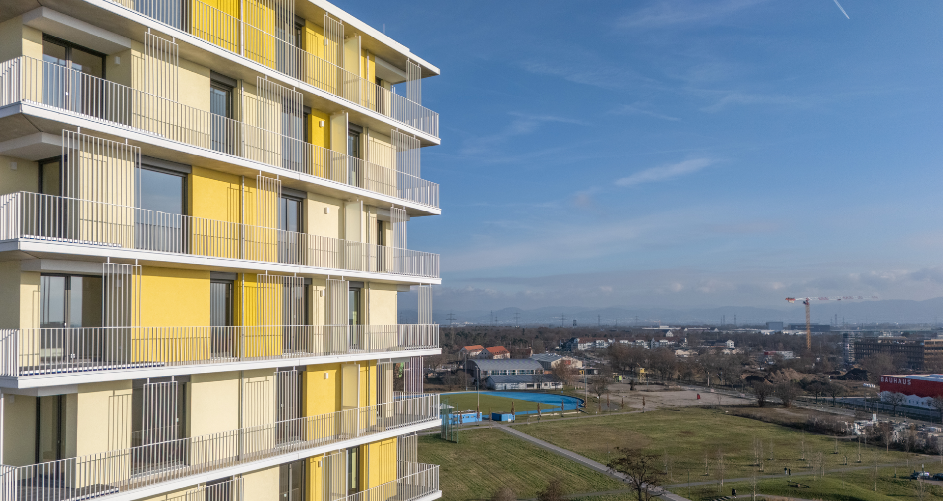 KI-basierte Bildbeschreibung: Auf der linken Seite ist ein modernes Wohngebäude mit gelben und beigen Paneelen und Balkonen zu sehen. Im Hintergrund befinden sich ein grasbewachsener Park, Bäume und entfernte Gebäude unter einem blauen Himmel mit leichten Wolken.