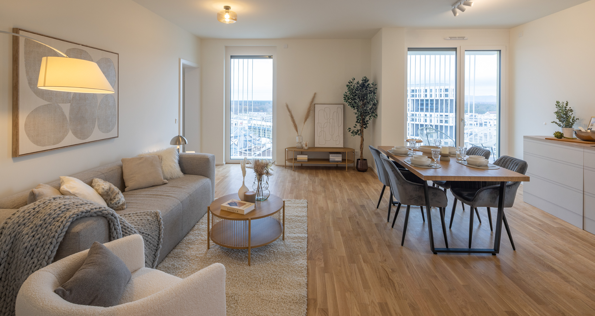 KI-basierte Bildbeschreibung: Moderner, offener Wohn- und Essbereich mit Holzfußboden, neutralen Möbeln, einem grauen Sofa, einem Sessel, einem Esstisch für sechs Personen, großen Fenstern mit Blick auf die Stadt und einer minimalistischen Einrichtung.