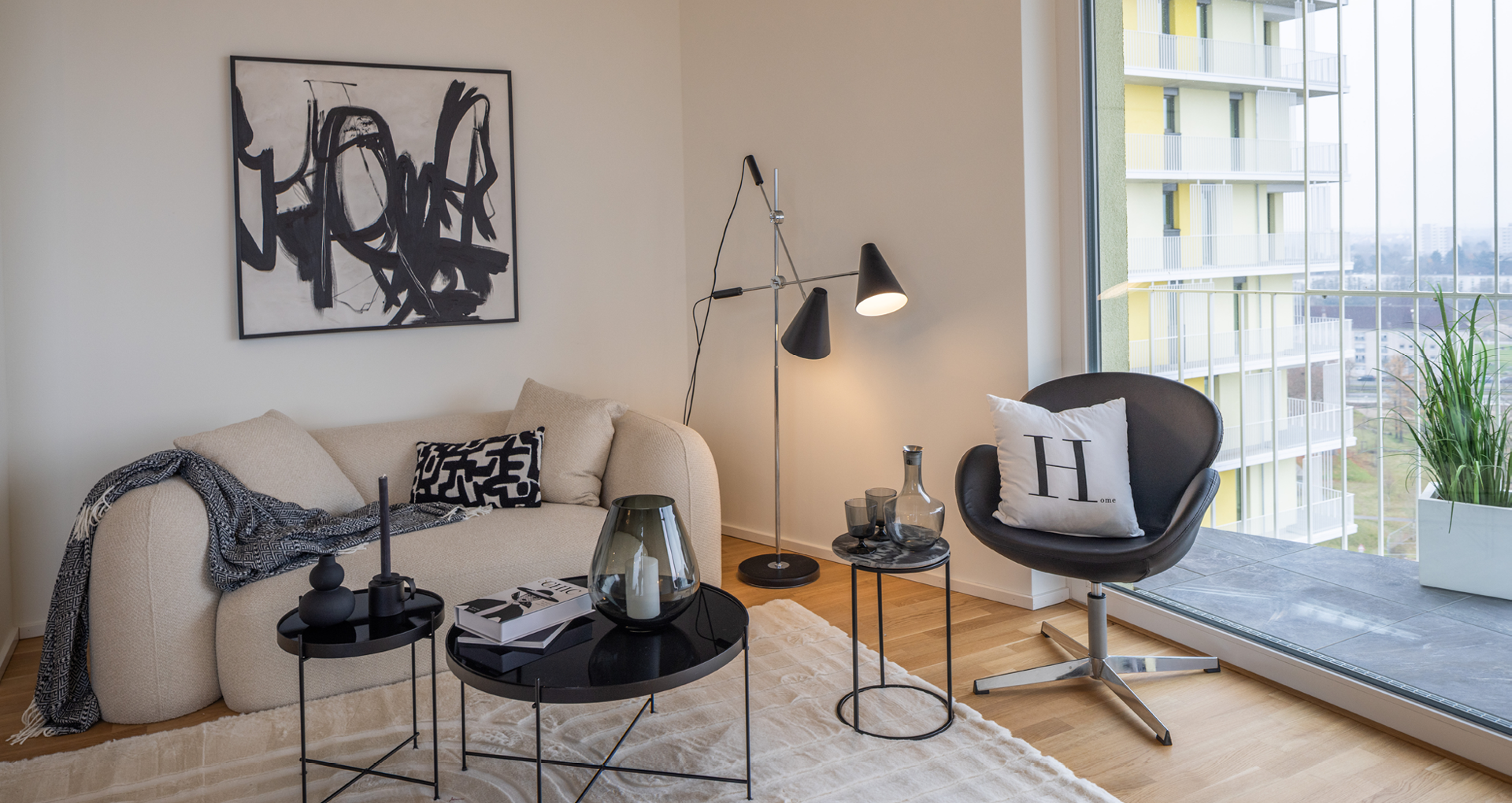 KI-basierte Bildbeschreibung: Ein modernes Wohnzimmer mit einem beigen Sofa, schwarz-weißen Kissen, abstrakter Wandkunst, schwarzen runden Couchtischen, einem schwarzen Stuhl mit einem Kissen, einer Stehlampe und einem großen Fenster mit Blick auf das Gebäude und die Stadt.