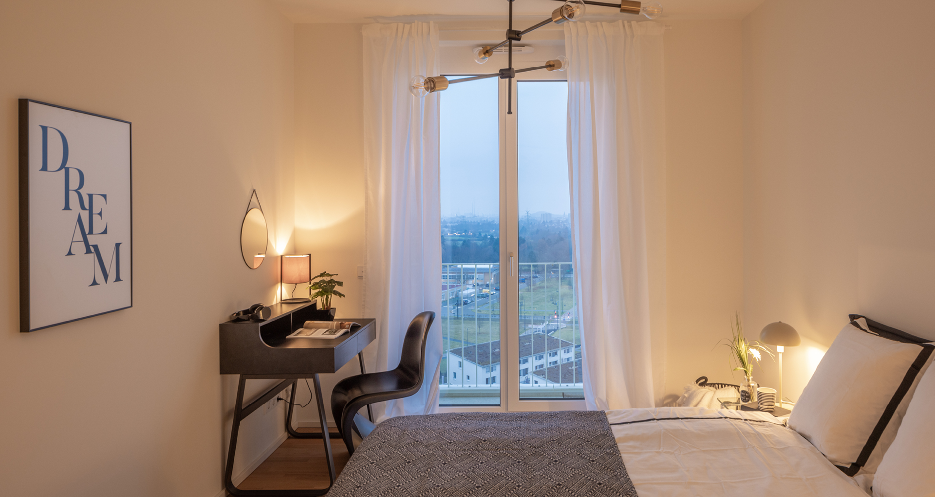 KI-basierte Bildbeschreibung: Ein gemütliches Schlafzimmer mit einem Bett, einem kleinen schwarzen Schreibtisch und Stuhl, einer Lampe, einer Pflanze und einem großen Fenster mit weißen Vorhängen mit Blick auf eine neblige Stadt. An der Wand hängt ein gerahmter "DREAM"-Druck.
