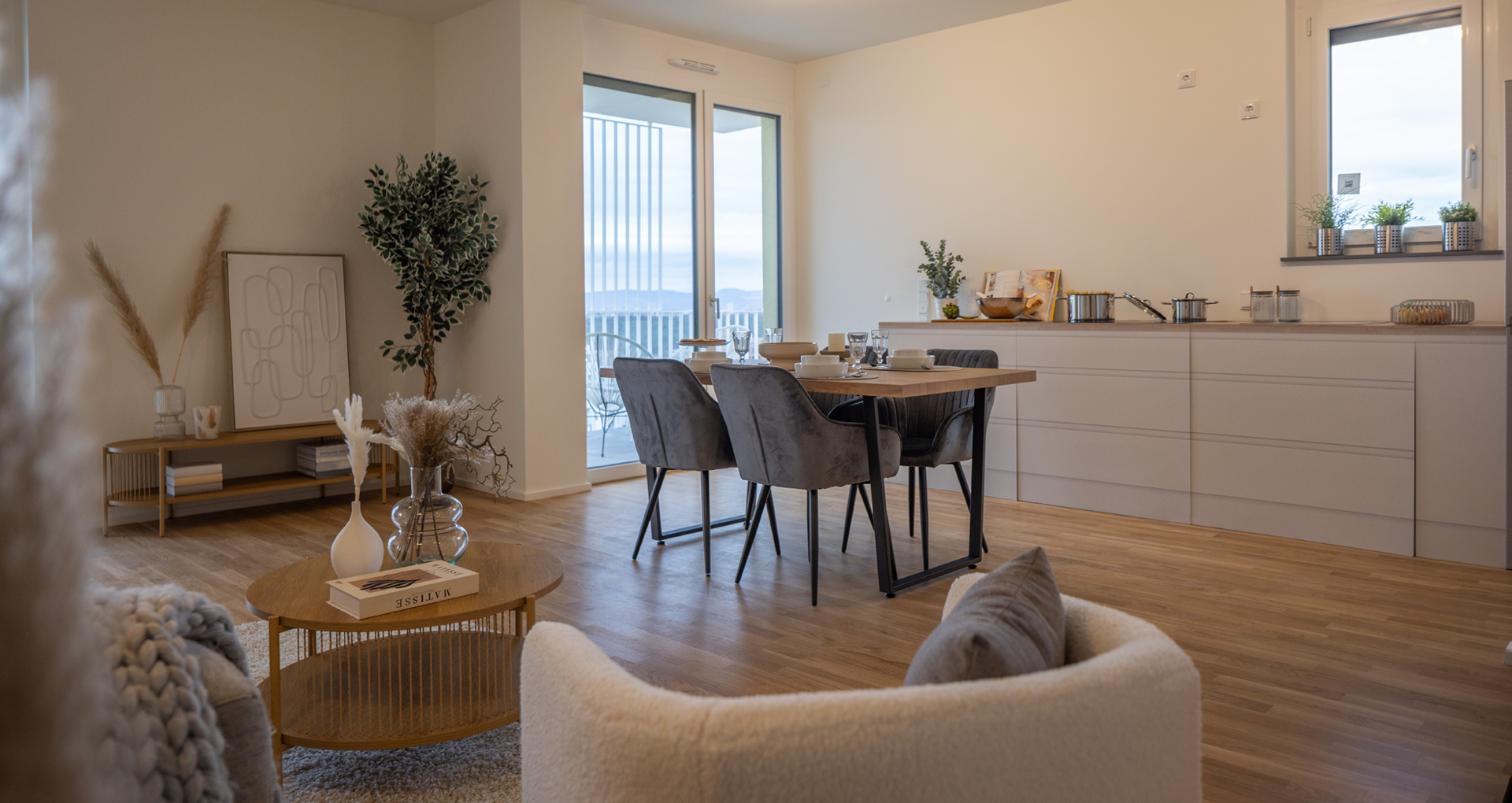 KI-basierte Bildbeschreibung: Ein moderner, offener Wohn- und Essbereich mit Holzböden, neutralen Möbeln, einem runden Couchtisch, einem gedeckten Esstisch und Küchenschränken. Große Fenster lassen natürliches Licht herein und bieten einen Blick nach draußen.