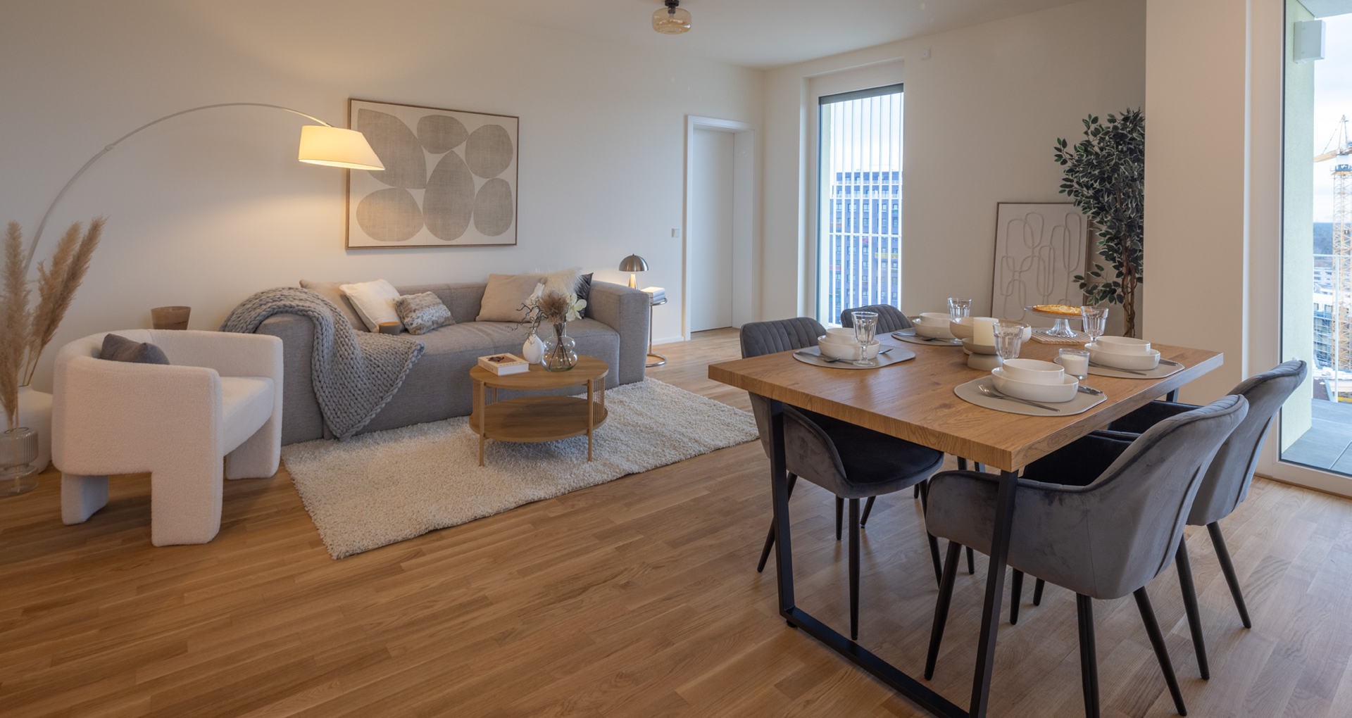 KI-basierte Bildbeschreibung: Moderner Wohn- und Essbereich mit einem grauen Sofa, Sessel und Teppich auf einem Holzboden. Esstisch für sechs Personen mit grauen Stühlen. Großes Fenster und natürliches Licht; neutrale Töne und minimalistisches Dekor in der gesamten Wohnung.