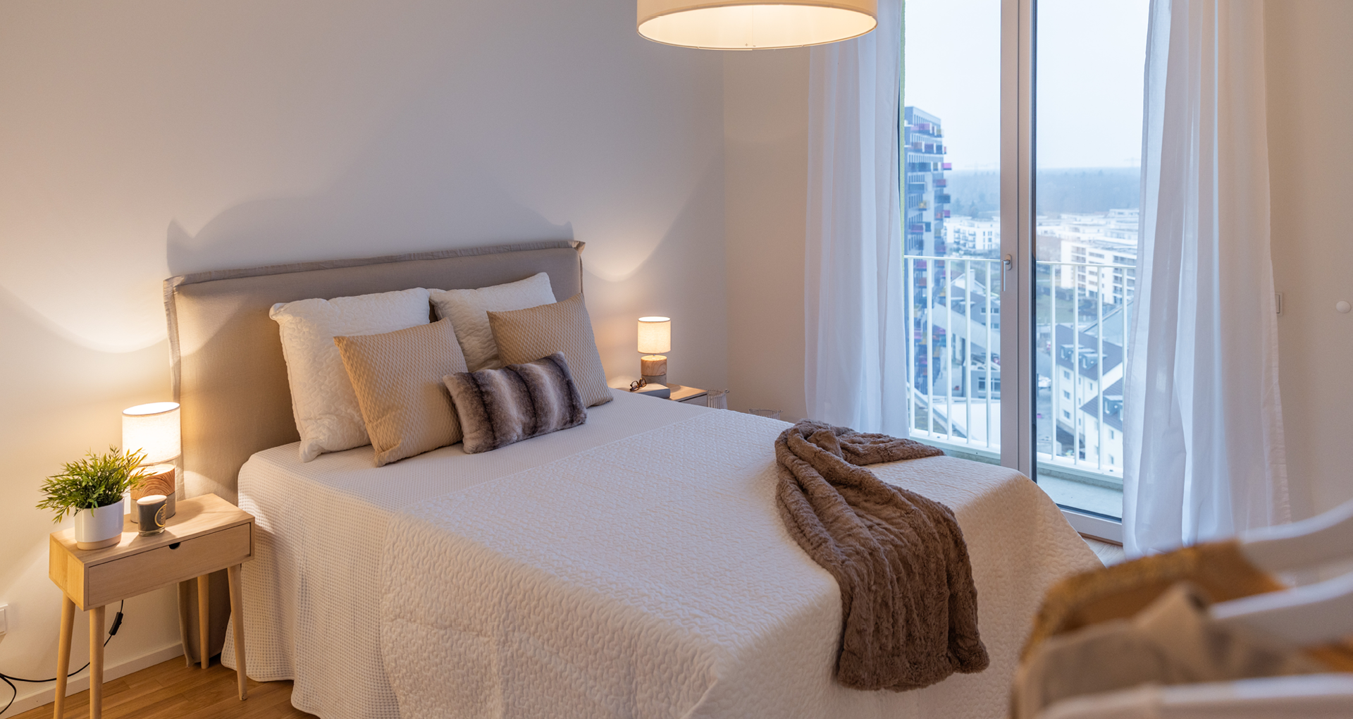 KI-basierte Bildbeschreibung: Ein modernes Schlafzimmer mit einem sauber gemachten Doppelbett, beiger und weißer Bettwäsche, dekorativen Kissen, einer braunen Überwurfdecke, zwei hölzernen Nachttischen mit Lampen und einem großen Fenster mit Blick auf die Gebäude der Stadt.