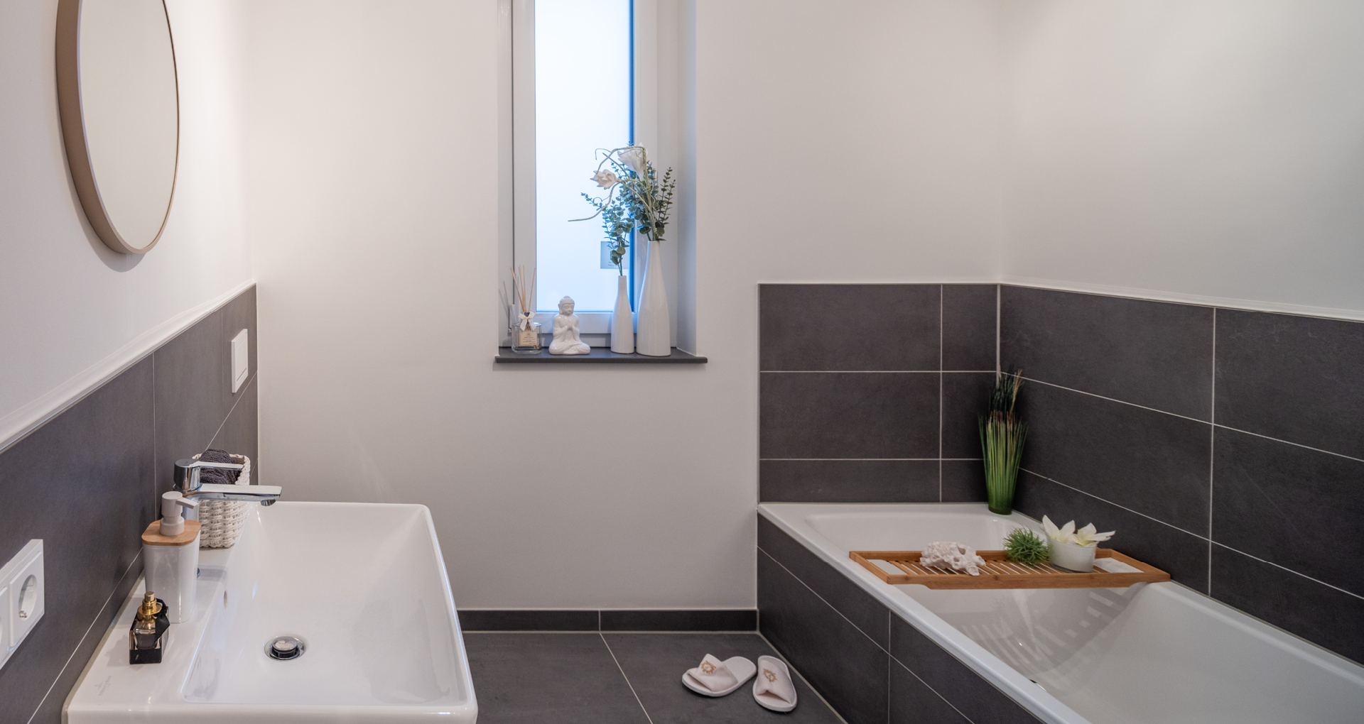 KI-basierte Bildbeschreibung: Modernes Badezimmer mit dunklen Fliesenwänden, weißem Waschbecken, Badewanne mit Holztablett, auf dem ein Handtuch und eine Pflanze stehen, einem kleinen Fenster mit Dekorationsgegenständen und weißen Hausschuhen auf dem Boden.